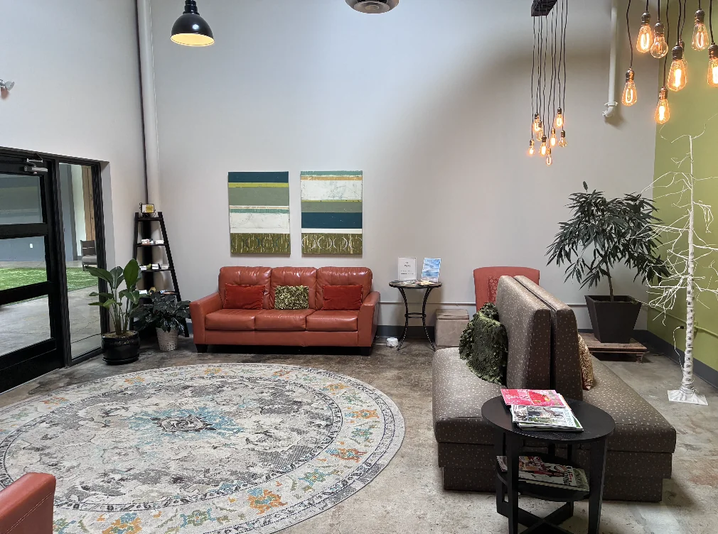 Sala de espera com assentos vermelhos e cinza, plantas, luminárias pendentes e um tapete redondo.
